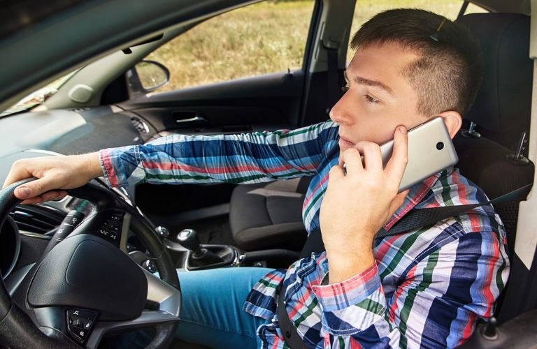 A man in checked shirt is talking on the phone while sitting on a car.